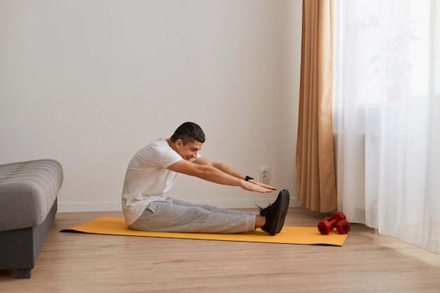 Plan intérieur d'un homme athlétique sportif portant des vêtements de sport assis sur un tapis de yoga par terre à la maison et faisant des étirements ayant une flexibilité d'entraînement d'entraînement