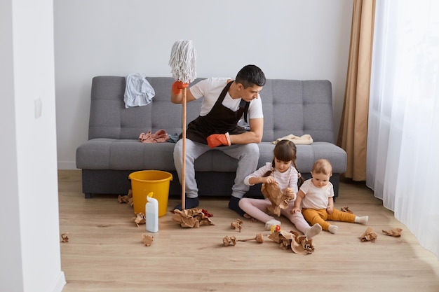 Plan intérieur d'un homme lavant le sol avec une vadrouille à la maison assis sur un canapé pour se reposer et parler avec sa fille qui joue par terre avec un père en papier regardant les enfants avec une expression sérieuse