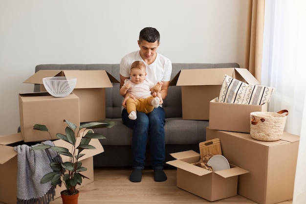 Plan intérieur d'un homme portant un t-shirt blanc assis sur un canapé avec sa petite fille lors d'un déménagement dans une nouvelle maison entourée de colis en carton avec des effets personnels