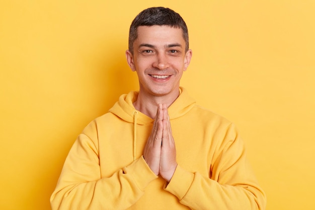 Plan intérieur d'un homme positif portant un sweat à capuche de style décontracté posant isolé sur fond jaune debout dans une pose de yoga et essayant de se détendre garde les paumes ensemble