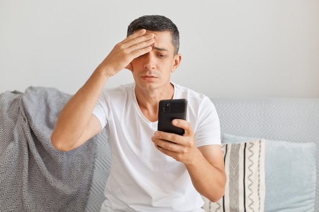 Plan intérieur d'un homme triste bouleversé portant un t-shirt blanc de style décontracté assis sur un canapé à l'aide d'un téléphone portable avec une expression de tristesse gardant la main sur la tête souffrant de maux de tête