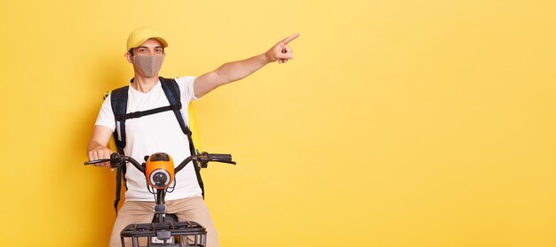 Plan intérieur d'un jeune adulte caucasien livreur à vélo portant une casquette de t-shirt blanc et un masque de protection livrant des commandes pendant la quarantaine pointant de côté montrant l'espace de copie pour la publicité