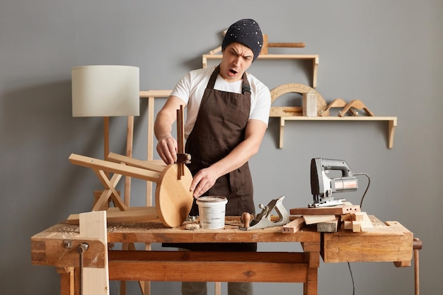 Plan intérieur d'un jeune charpentier choqué et étonné portant un t-shirt blanc, une casquette noire et un tablier marron travaillant menuisier sur son lieu de travail ayant une erreur en faisant une chaise en bois