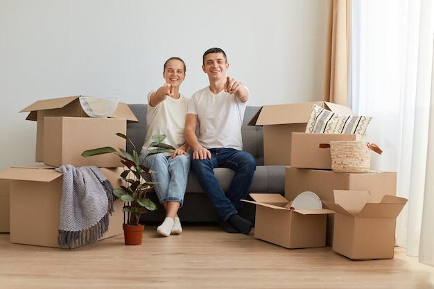 Plan intérieur d'un jeune couple heureux et excité assis sur un canapé entouré de boîtes en carton avec des effets personnels pointant vers la caméra vous choisissant pour l'achat d'un nouvel appartement hypothécaire