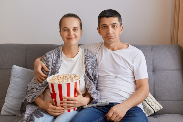 Plan intérieur d'un jeune couple regardant la télévision à la maison assis sur la toux étreignant sa femme enveloppée dans un plaid et tenant un seau avec une famille de pop-corn passant du temps ensemble