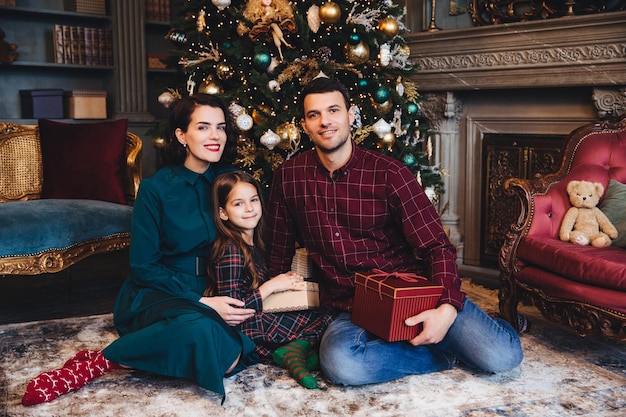 Plan intérieur d'une jeune famille qui a de bonnes relations s'asseoir ensemble sur le sol près d'un arbre du Nouvel An décoré tenir présent se féliciter avec le concept du Nouvel An et des vacances de Noël à venir