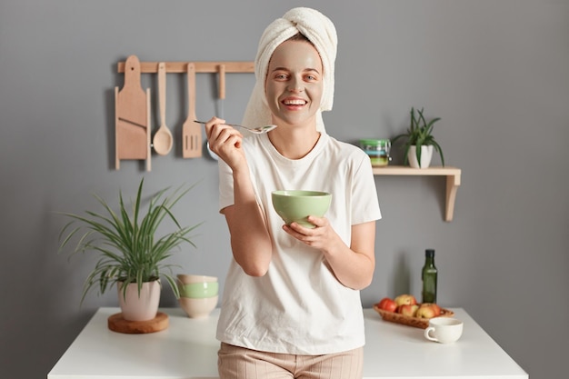 Plan intérieur d'une jeune femme adulte caucasienne enveloppée d'une serviette blanche et vêtue d'un t-shirt en train de manger son petit-déjeuner et de faire des procédures de soins de la peau debout dans la cuisine à la maison