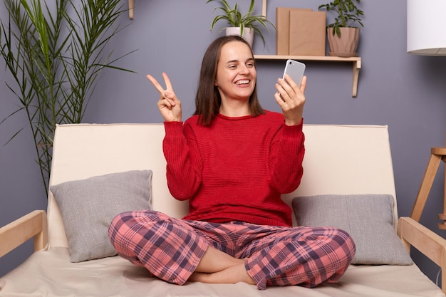 Plan intérieur d'une jeune femme adulte satisfaite portant un pull rouge et un pantalon à carreaux assis sur un canapé à l'intérieur de la maison tenant un téléphone intelligent ayant un appel vidéo ou faisant un selfie souriant et montrant le signe v