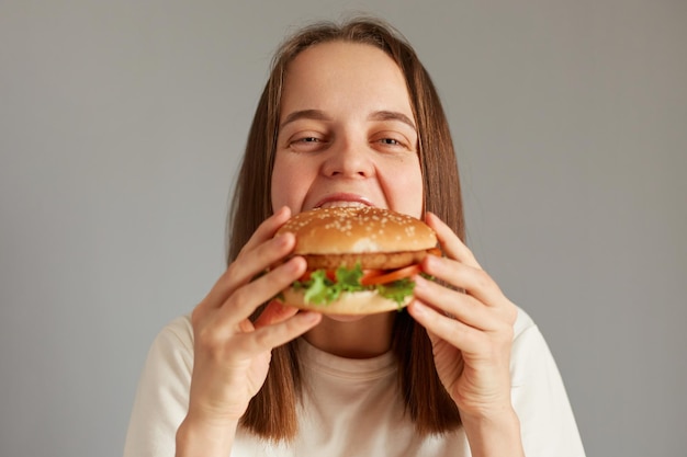 Plan intérieur d'une jeune femme adulte séduisante appréciant la bouchée d'un délicieux hamburger appétissant isolé sur fond gris mangeant un hamburger savoureux regardant la caméra avec un visage satisfait