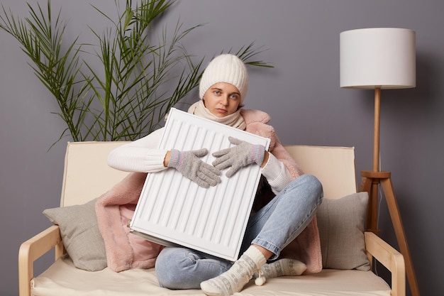 Plan intérieur d'une jeune femme adulte triste en manteau et chapeau assis dans un salon froid tenant un radiateur électrique luttant contre le froid glacial à la maison regardant la caméra exprimant sa tristesse