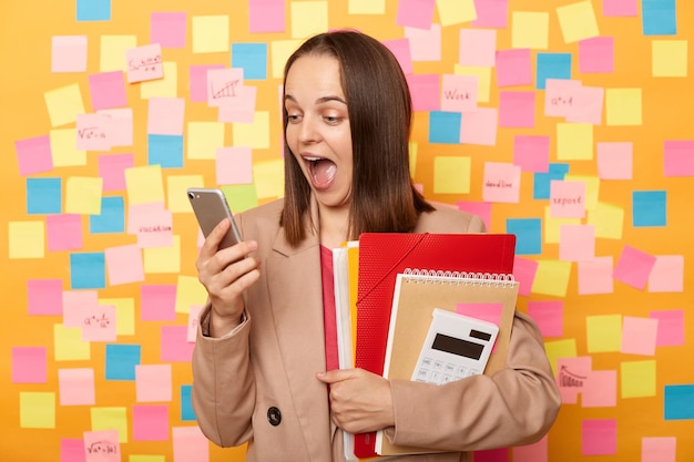 Plan intérieur d'une jeune femme étonnée extrêmement excitée tenant un dossier papier de cahiers et une calculatrice à l'aide d'un téléphone intelligent lisant les dernières nouvelles posant contre un mur jaune avec des autocollants colorés