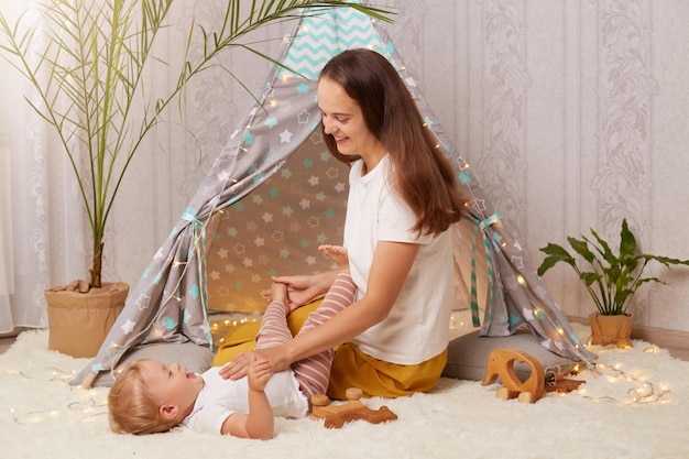 Plan intérieur d'une jeune mère jouant avec sa petite fille sur le sol près d'un wigwam pour enfants exprimant des émotions positives passant du temps avec un enfant mignon dans une tente peetee à la maison