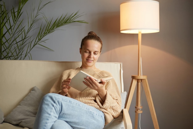 Plan intérieur d'une jolie femme aux cheveux bruns ravie portant un pull beige et un jean assis sur la toux avec un livre dans les mains lisant un roman intéressant ou de beaux poèmes