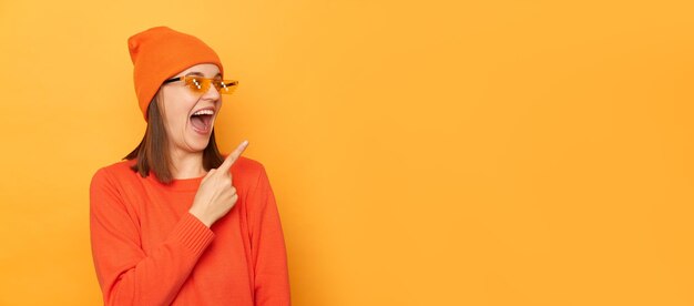 Plan intérieur d'une jolie femme heureuse portant un bonnet de pull orange et des lunettes dans le style minecraft indiquant loin dans la zone de publicité debout isolé sur fond jaune