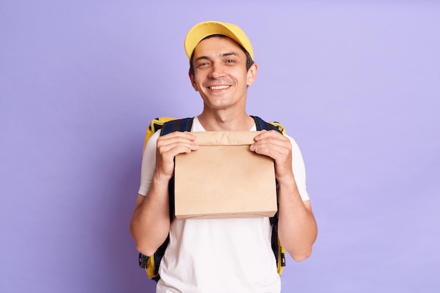 Plan intérieur d'un livreur souriant et satisfait tenant un paquet de papier avec de la nourriture isolé sur fond violet regardant la caméra avec un sourire à pleines dents apportant l'ordre au client