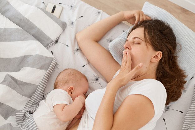 Plan intérieur d'une mère de femme fatiguée et endormie aux cheveux noirs couvrant la bouche, une femme portant un t-shirt décontracté blanc, allaitant son bébé, une maternité heureuse.