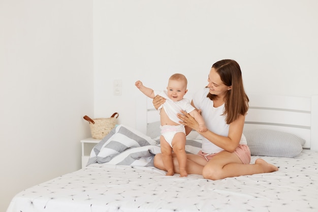 Plan intérieur d'une mère tenant son bébé dans la chambre alors qu'elle était assise sur le lit, une femme aux cheveux noirs portant un t-shirt décontracté blanc, un tout-petit debout avec l'aide de maman et riant.