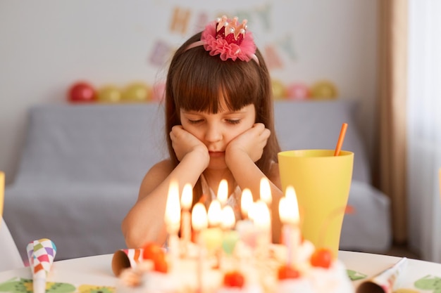 Plan intérieur d'une petite fille charmante célébrant son anniversaire seule, attendant des invités, assise à table avec un gâteau lumineux avec des bougies, gardant les mains sur son visage, exprimant sa tristesse et sa tristesse.