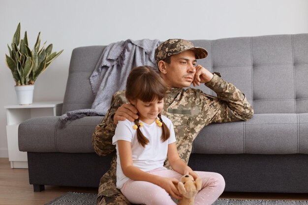 Plan intérieur d'un soldat caucasien pensif portant un uniforme de camouflage rentrant chez lui après l'armée, passant du temps avec sa petite fille et détournant les yeux avec une expression faciale réfléchie.