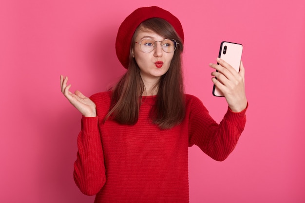 Plan intérieur d'une superbe femme de race blanche faisant un geste de baiser tout en prenant une photo d'elle-même. Portrait de romantique de dame aux cheveux noirs robes béret et pull, rend selfie sur rose