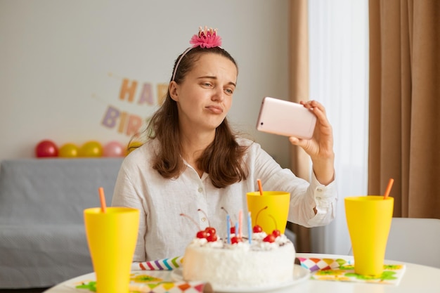 Plan intérieur d'un triste contrarié portant des vêtements blancs assis à table avec un gâteau et une boisson, ayant un appel vidéo ou diffusant en direct, sent que les invités tristes ne viennent pas.