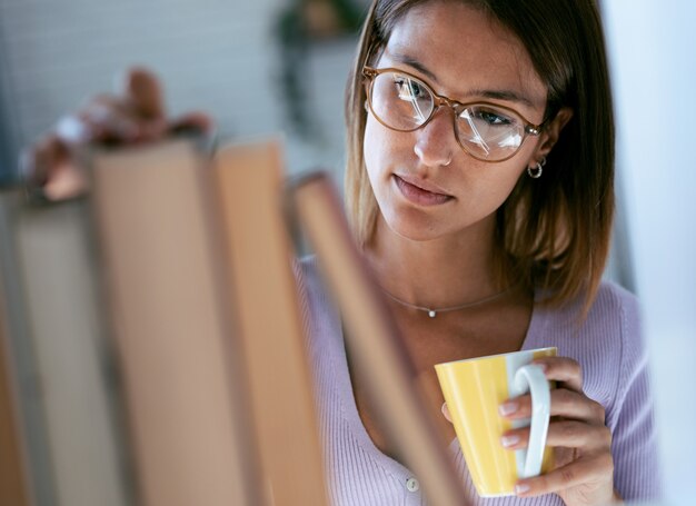 Plan d'une jeune femme intelligente buvant une tasse de café tout en cherchant un livre dans la bibliothèque à la maison.