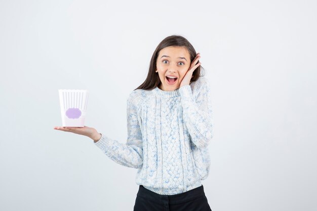 Plan d'une jeune fille souriante en pull tenant une boîte de pop-corn.