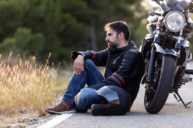 Plan d'un jeune homme assis sur la route, appuyé sur sa moto en attendant que l'assistance routière répare la moto.