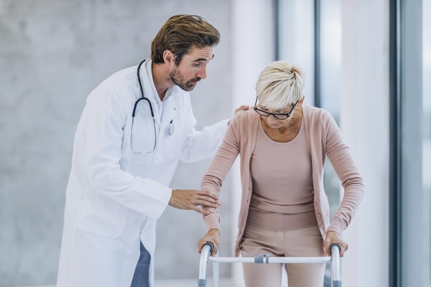 Plan d'un jeune médecin aidant une patiente âgée à marcher avec son déambulateur dans le couloir de l'hôpital.