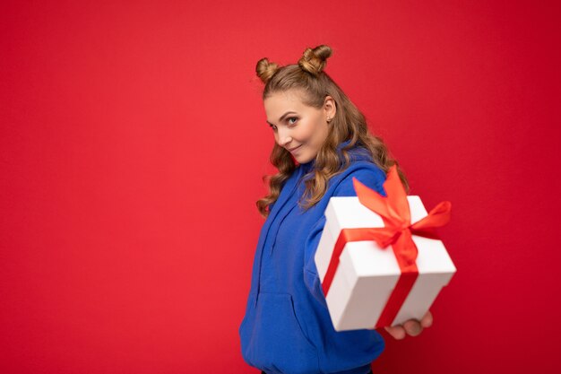 Plan d'une jolie jeune femme blonde heureuse isolée sur un mur de fond rouge portant un sweat à capuche tendance bleu tenant une boîte-cadeau et regardant la caméra. Copier l'espace, maquette