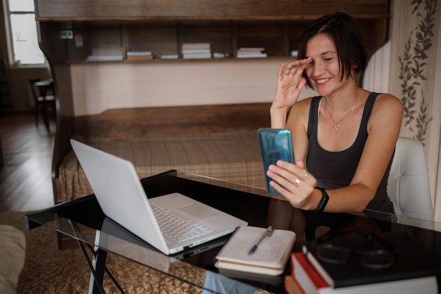Plan d'une jolie jeune femme utilisant son téléphone portable tout en travaillant avec un ordinateur portable assis à la maison
