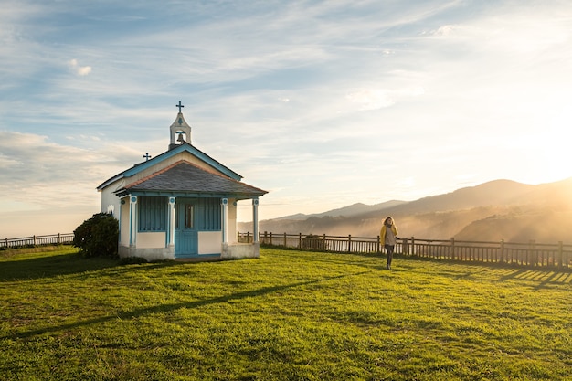 Plan large d'une femme marchant à côté d'une petite chapelle sur une falaise pendant le lever du soleil