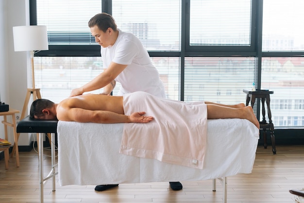 Plan large latéral d'un masseur masculin professionnel avec de fortes mains tatouées massant le dos et l'épaule d'un sportif musclé allongé sur le ventre à la table de massage Concept de mode de vie sain et de repos