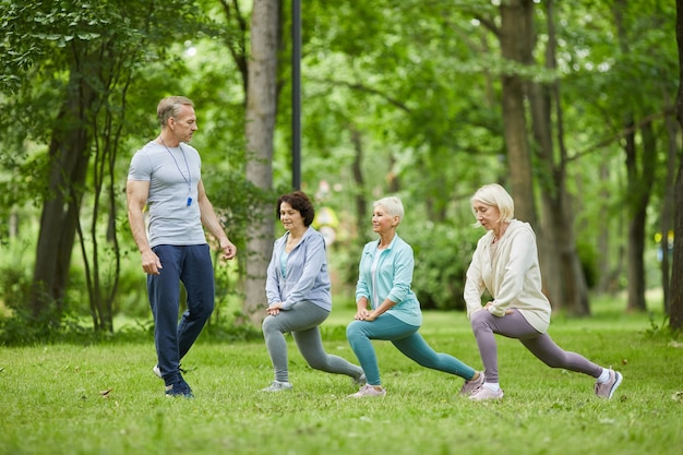 Plan large de trois belles femmes âgées ayant une séance d'entraînement dans un parc de la ville avec un entraîneur qui les regarde