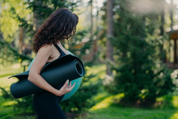 Plan latéral d'une femme brune porte une balle en forme et karemat enroulé va faire de l'exercice de fitness se détourne ne regarde pas les poses de caméra sur fond de nature verte floue