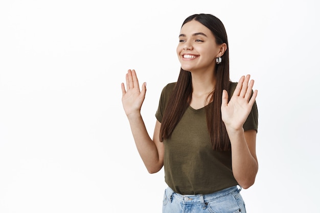 Plan latéral d'une fille souriante et joyeuse levant les mains, l'air amical et heureux de côté dans l'espace de copie de gauche, renonçant à quelqu'un pour saluer ou dire bonjour, debout contre un mur blanc