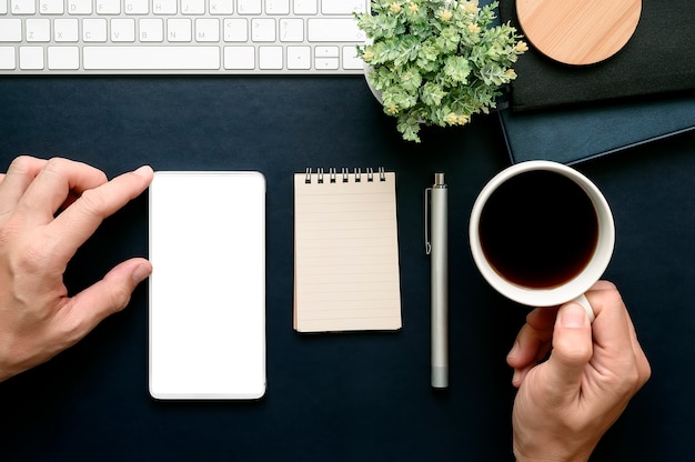 Plan de la main d&#39;un homme touchant l&#39;écran d&#39;un smartphone et tenant une tasse de café tout en travaillant au bureau.