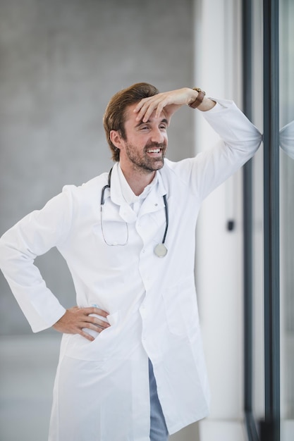 Plan d'un médecin souriant debout et pensif regardant par la fenêtre tout en faisant une pause rapide dans un couloir d'hôpital.