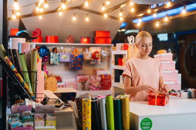 Plan moyen d'une jeune vendeuse tenant et utilisant un ruban doré pour nouer un nœud pour une boîte cadeau enveloppée de rouge...