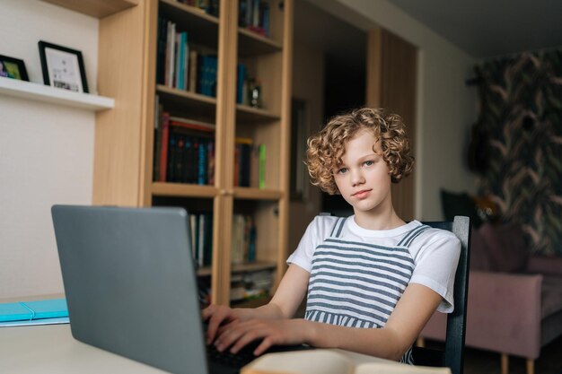 Plan moyen d'une jolie petite écolière bouclée assise à table en regardant la caméra faire ses devoirs à l'aide d'un ordinateur portable et d'un manuel papier