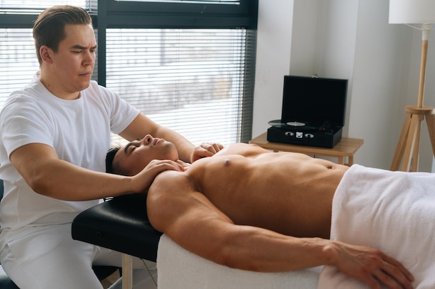 Plan moyen d'un masseur masculin professionnel avec des mains fortes massant l'épaule d'un athlète musclé sur un traitement de spa, allongé sur le dos dans une table de massage sur fond de fenêtre.