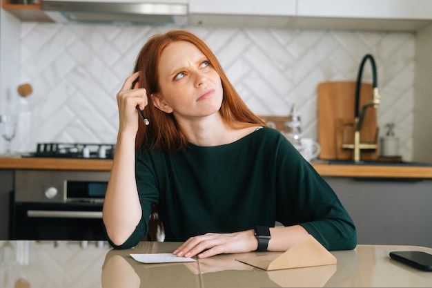 Plan moyen de la réflexion d'une jeune femme écrivant une lettre manuscrite assise à table avec une enveloppe en