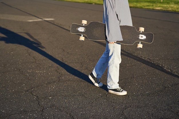 Plan recadré du corps d'une adolescente tenant un longboard de croiseur à la main marchant dans des baskets sur la route en jean