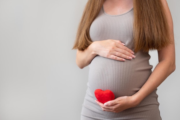 Plan recadré d'une femme enceinte portant une robe moulante tenant un coeur rouge doux sur fond gris Grossesse amour soins de santé et concept de maternité Idée de problèmes de cardiologie ou de maladieCopier l'espace