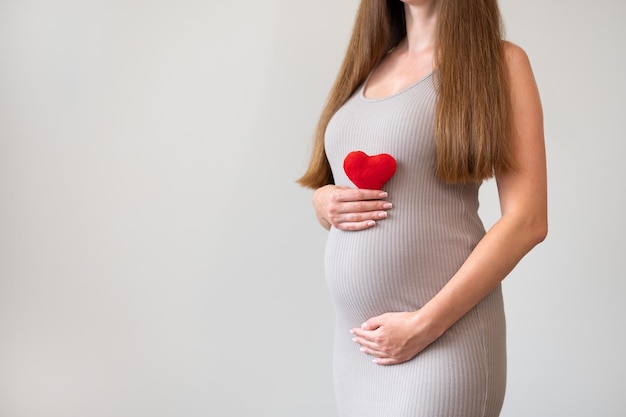 Plan recadré d'une femme enceinte portant une robe moulante tenant un coeur rouge doux sur fond gris Grossesse amour soins de santé et concept de maternité Idée de problèmes de cardiologie ou de maladieCopier l'espace