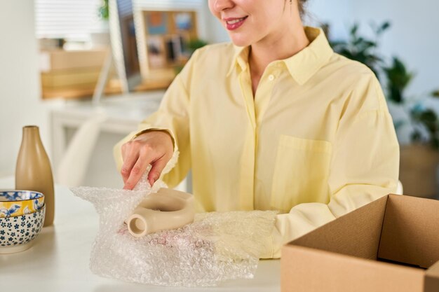 Plan recadré d'une jeune femme souriante enveloppant un vase en céramique dans de la cellophane protectrice avant de le mettre dans une boîte et de l'emballer