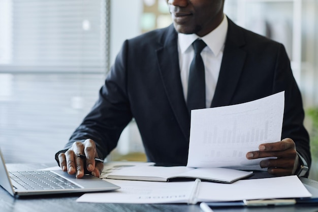 Plan recadré d'un jeune homme d'affaires noir portant un costume à l'aide d'un ordinateur portable sur le lieu de travail au bureau et tenant