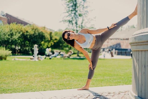 Plan sur toute la longueur de la femme brune étire les jambes montre une bonne flexibilité ne se divise habillé en lunettes de soleil de vêtements de sport pose à l'extérieur mène un mode de vie actif. Concept de sport et d'aérobic de personnes.