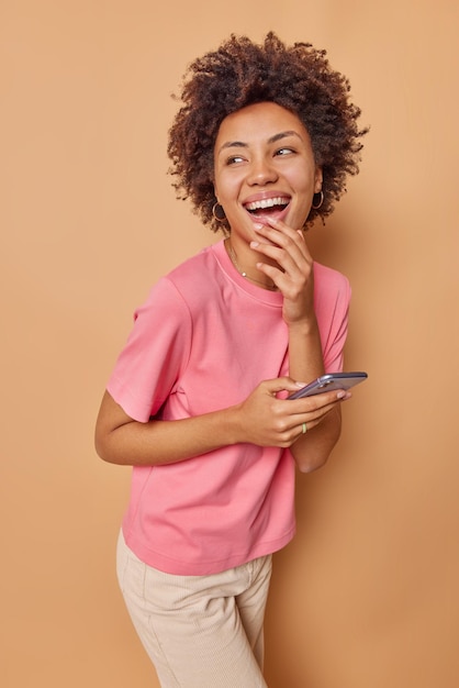 Plan vertical d'une adolescente optimiste et insouciante à l'expression positive portant des vêtements décontractés et tenant un téléphone portable pour discuter en ligne isolé sur beige. Émotions de technologie de personnes