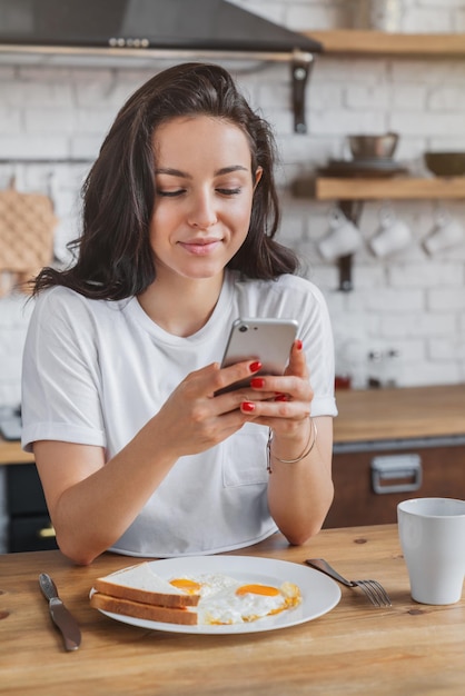 Plan vertical d'une belle jeune femme utilisant un téléphone portable tout en prenant son petit déjeuner dans une cuisine moderne